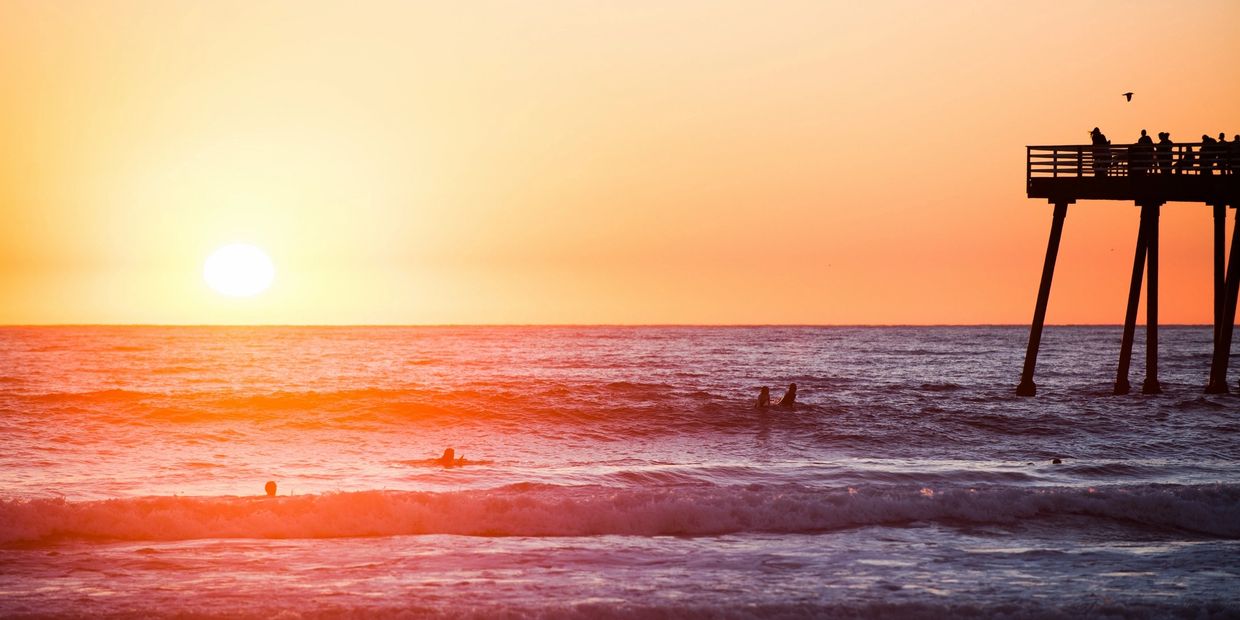 Sunset over the ocean with surfers and a pier.