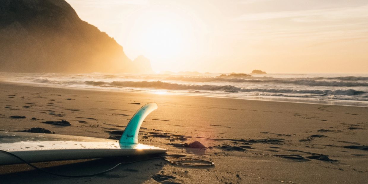 Surfboard resting on a sandy beach at sunrise with waves and cliffs in the background.