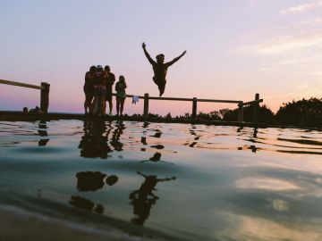 Person jumping off a dock into water during sunset with friends nearby.