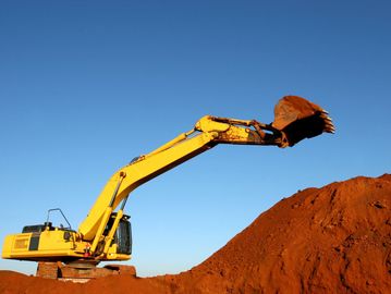 Yellow excavator digging a large pile of red soil under clear blue sky.