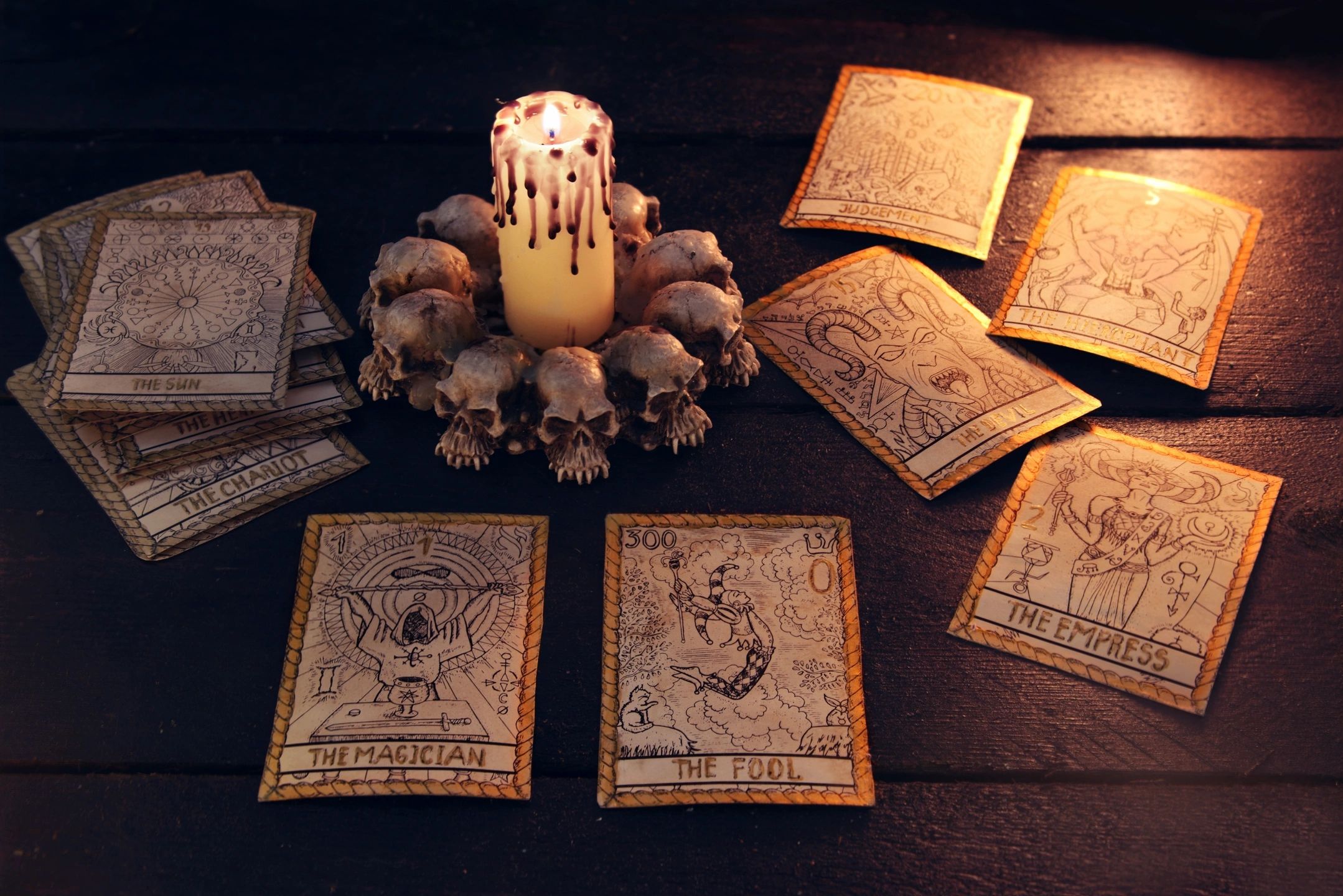 A lit pillar candle sits within a circle of skulls on a wooden table surrounded by tarot cards