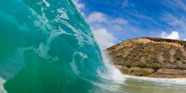 A powerful ocean wave curls near a rocky hill under a blue sky.