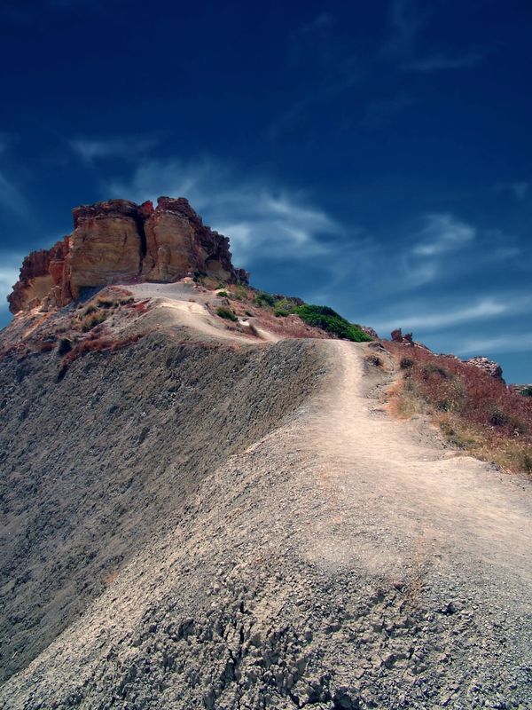 A narrow dirt path winding up toward a rocky peak under a vivid blue sky with light clouds.