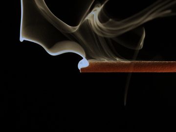 Close-up of incense stick burning with smoke curling upwards against black background.