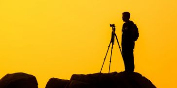 Silhouette of a photographer with a tripod on a rocky hill during sunset.