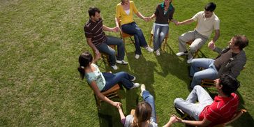 A group of people sitting in a circle on chairs holding hands outdoors.