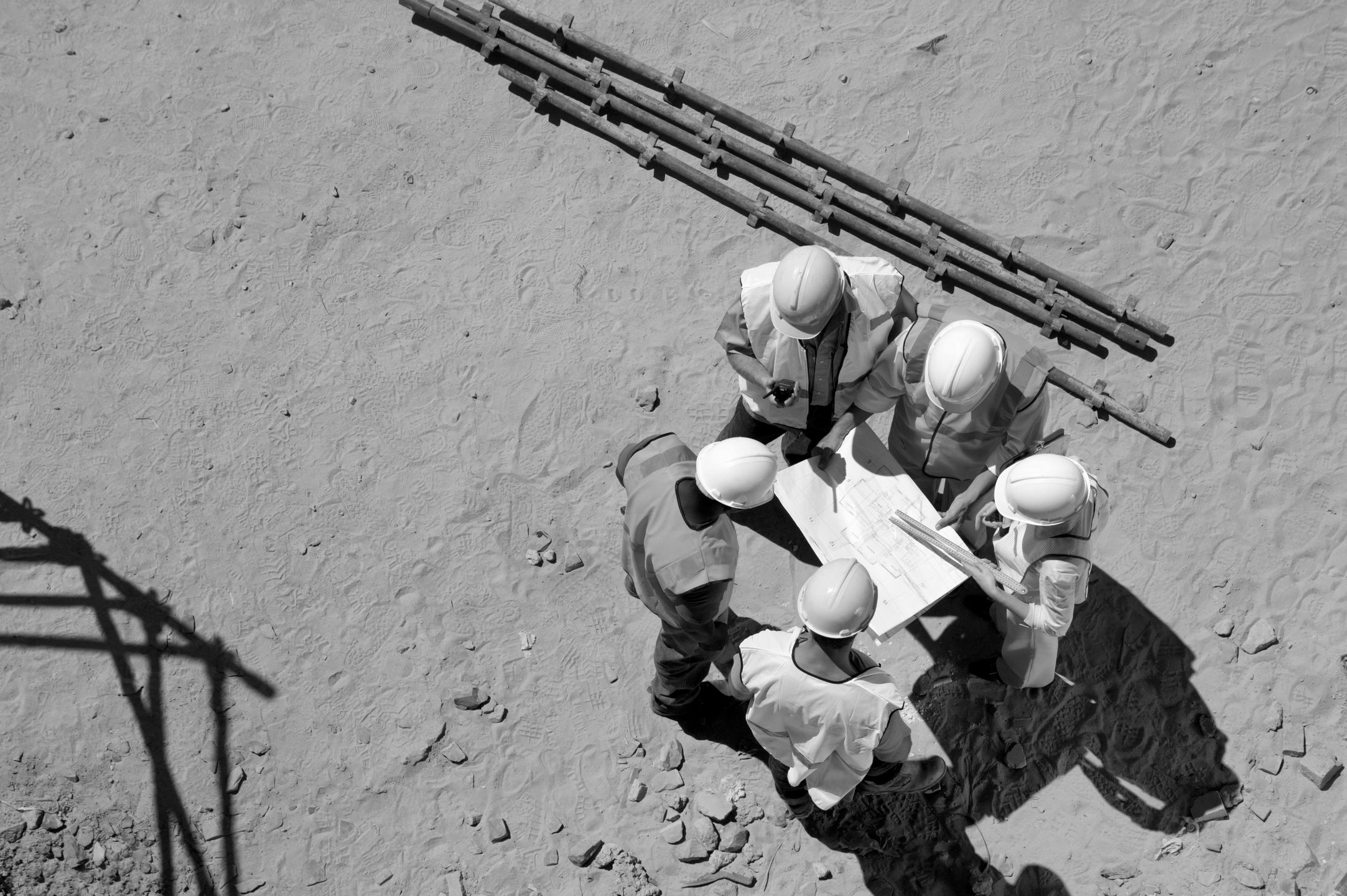 Construction workers reviewing plans at a sandy site with metal pipes nearby.