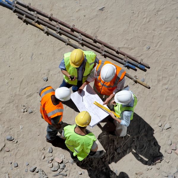 Construction workers reviewing plans on a sandy site with equipment nearby.