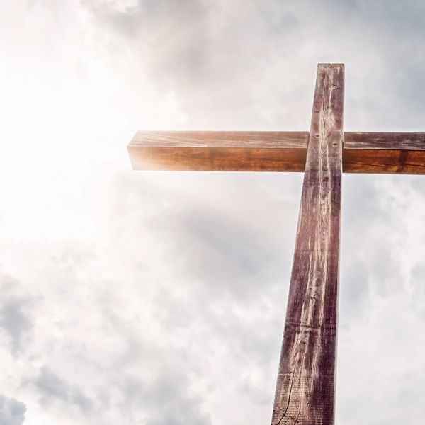 A wooden cross stands tall against a cloudy sky with sunlight.