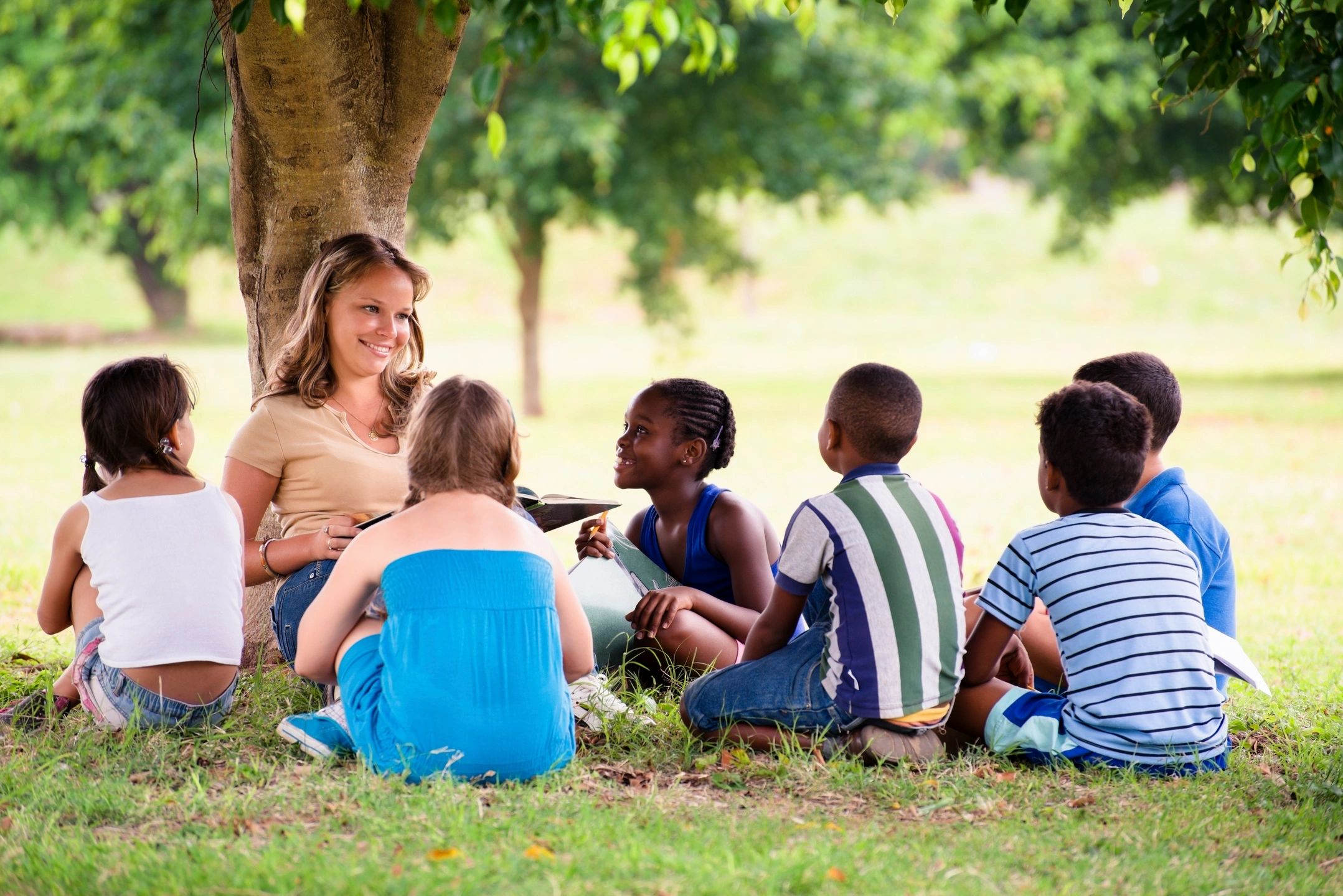 Woman teacher sitting at base of tree reading to 6 children on a summer day