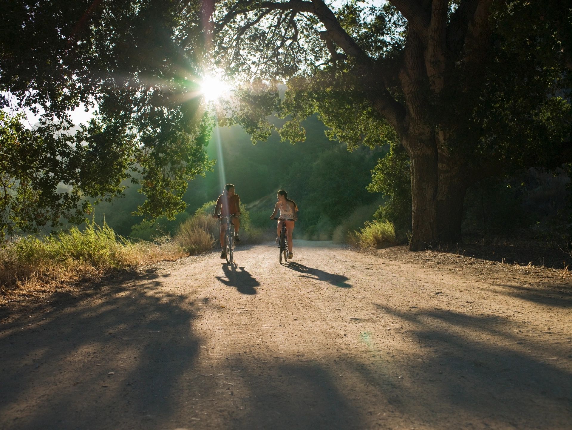 Man and woman riding pedal bikes down dirt road in wooded area on sunny day