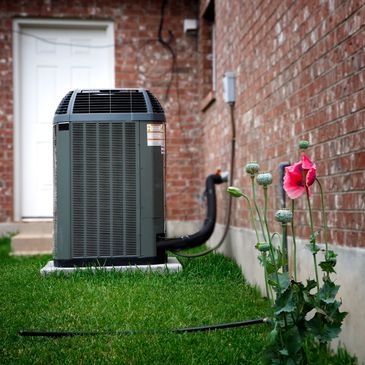 Outdoor air conditioning unit beside a brick house wall with flowers nearby.