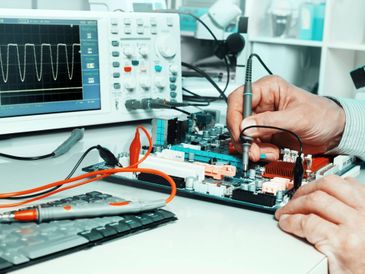 Hands testing an electronic circuit board with an oscilloscope in the background.