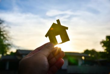 Hand holding a paper house cutout against the sunlit sky.