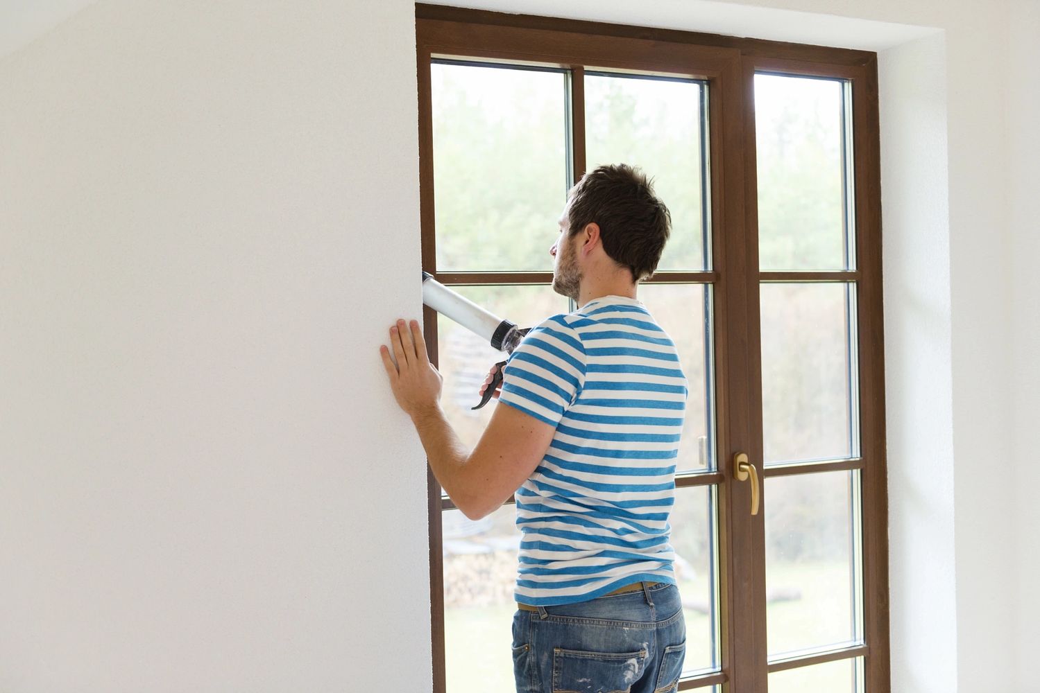 Man applying sealant around a window frame indoors.