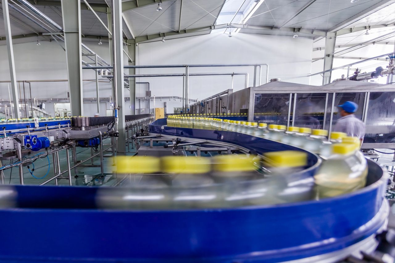 The factory assembly line moves forward with plastic bottles with yellow caps, lined up, with liquid in them, as one worker walks by.