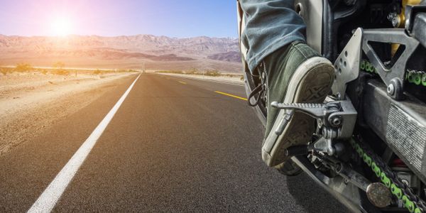 Rider's foot on motorcycle pedal on an open desert road at sunset.