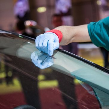 A human, wearing blue latex gloves, using a razor blade to clean off repair resin on a windshield. 
