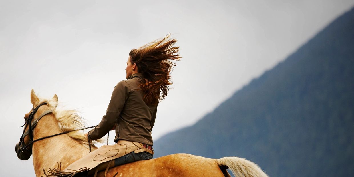 A woman with flowing hair rides a light brown horse against a mountainous backdrop.