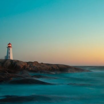 white and red lighthouse on the top of a cliff overlooking water with a subtle sunset in the backgro