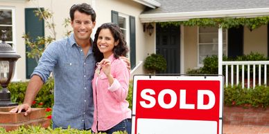 A man and woman are standing in front of their house with a sold sign.