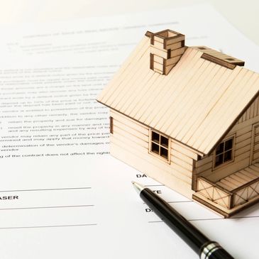 Small wooden model of a home laying on top of a paper contract with a pen laying next to it.