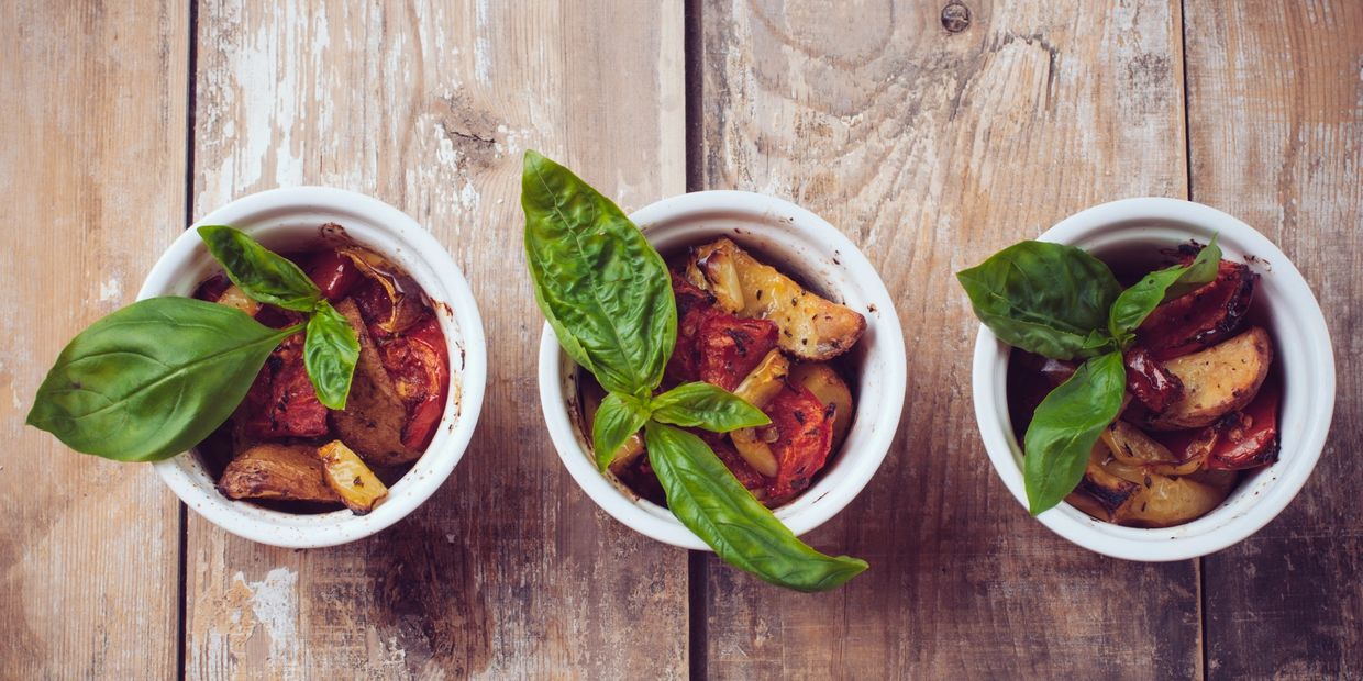 Three small bowls with roasted vegetables and fresh basil leaves on wooden table.