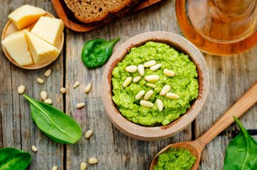 Bowl of green pesto topped with pine nuts surrounded by spinach and cheese.