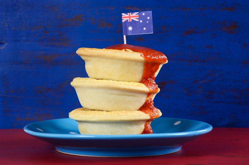3 Aussie meat pies stacked on top with red sauce running down side, mini Australia flag  on top
