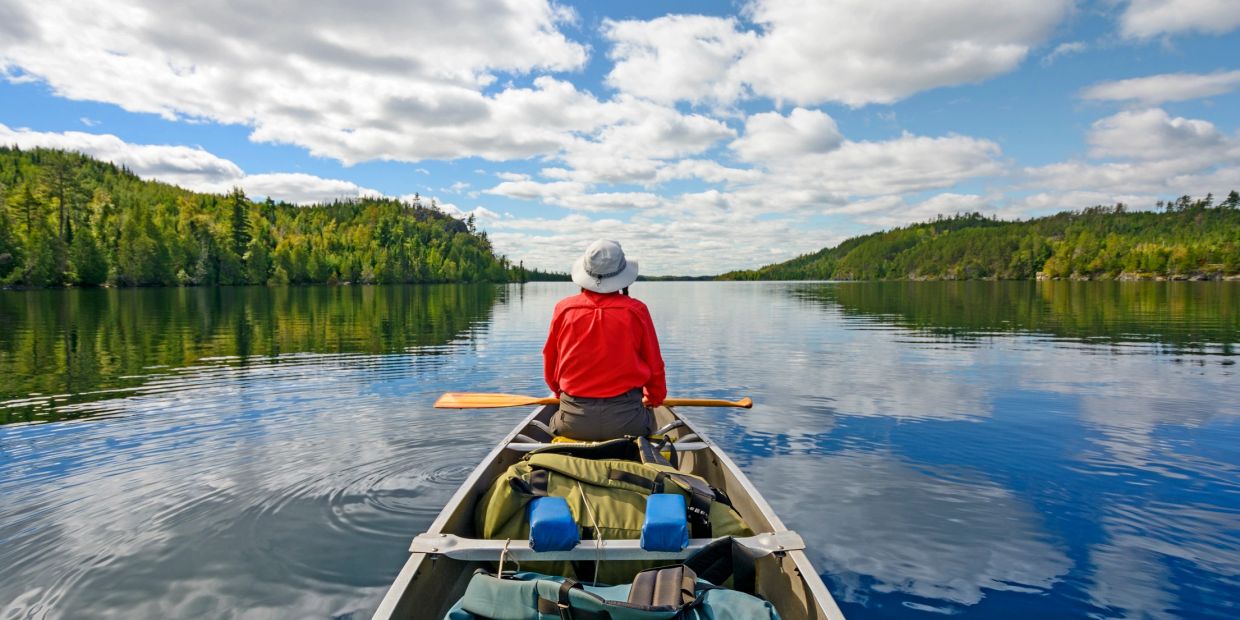 Person in red paddling a canoe on a calm lake surrounded by forested hills under a partly cloudy sky.