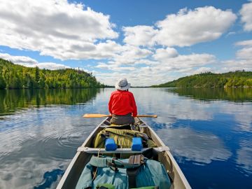 Person in red jacket paddling a canoe on a calm lake surrounded by green forests.