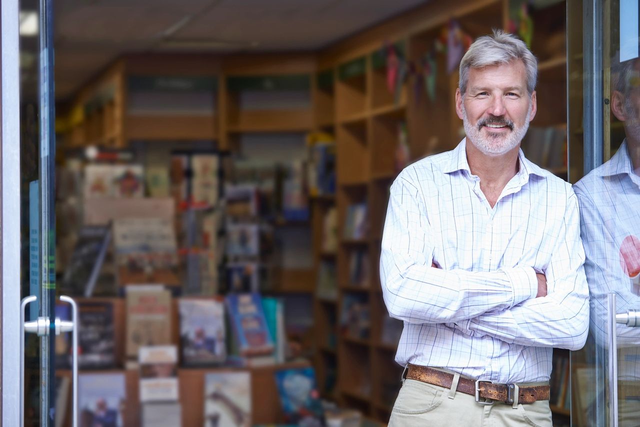 Owner stands, leaning in the entryway of his business smiling.