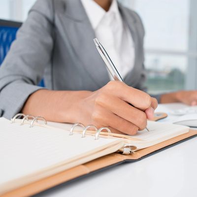 A person in business attire writing in a notebook with a pen.