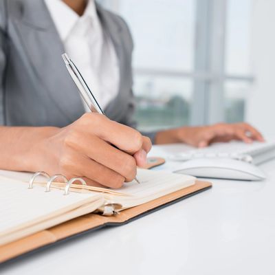 Person in business attire writing in a notebook with a keyboard nearby.