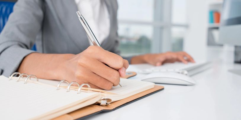 Person in business attire writing in a notebook at a desk.
