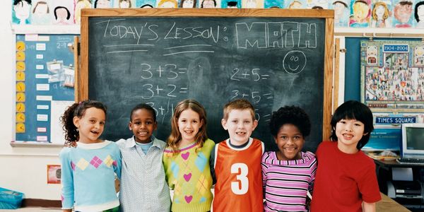 Children in a classroom standing in front of a chalk board smiling and hugging each other. 