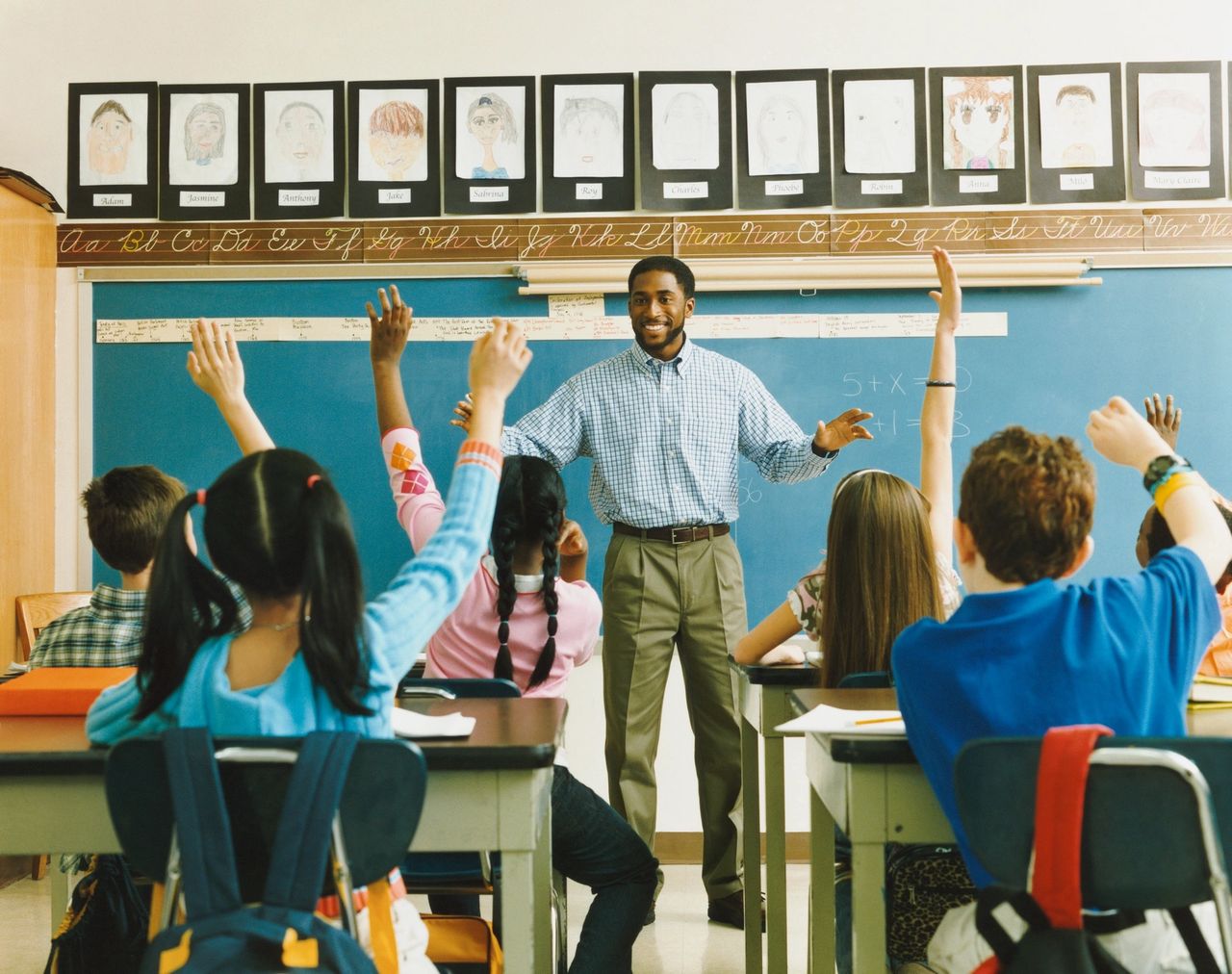 A teacher stands in front of the class with the chalk board behind him smiling as students eagerly raise their hands.