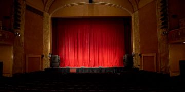 Dark theater with closed red curtains and empty seats.