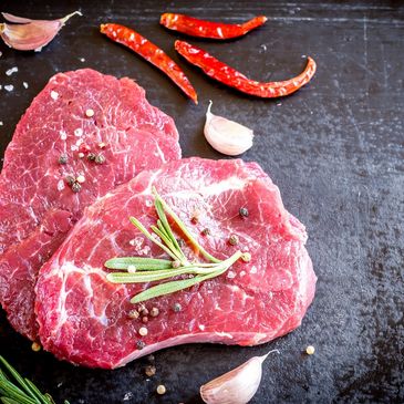 Raw beef steaks with rosemary, garlic, and chili peppers on a dark surface.