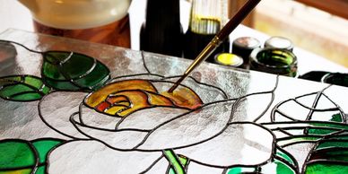 close up pic of a paintbrush filling in the yellow of a rose part of a stained glass window. 