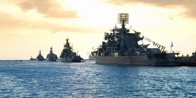 A fleet of navy warships lined up on calm water at sunset.