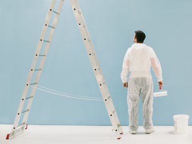 Painter in white suit looking at a blue wall with ladder and paint bucket.