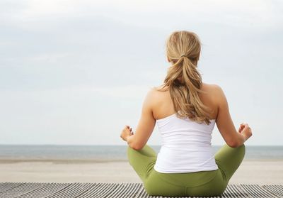 A woman sitting in a cross-legged position meditating and looking out over a cloudy beach. 