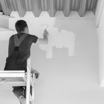 Man painting a wall white while standing on scaffolding.