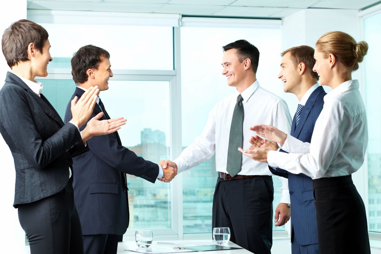 Business partners smile and shake hands as others clap their hands.