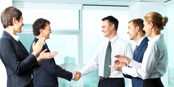 5 individuals in a business meeting, all smiling, with two of them shaking hands. 