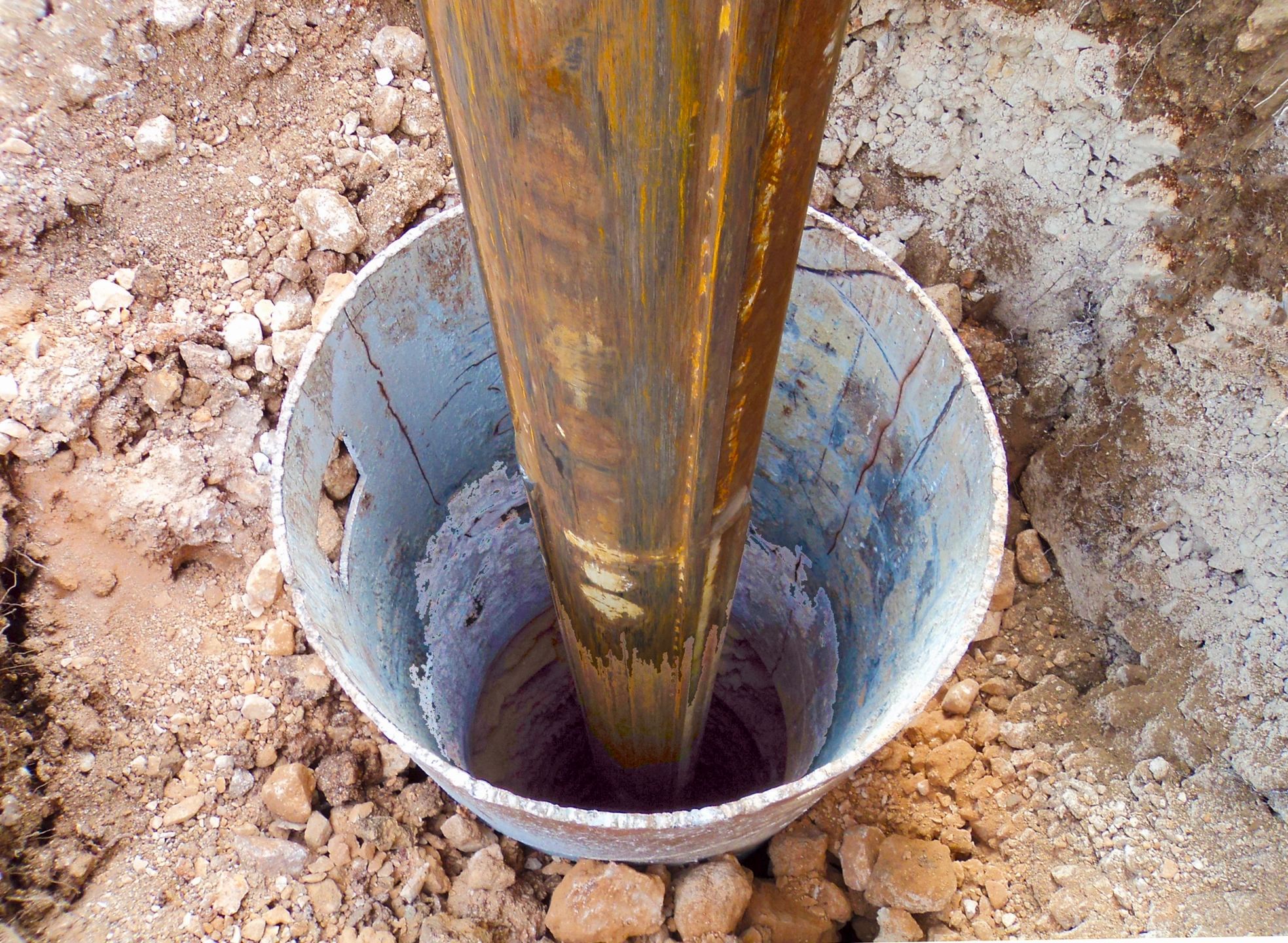 Rusty metal pipe inside a concrete cylinder surrounded by rocky soil.