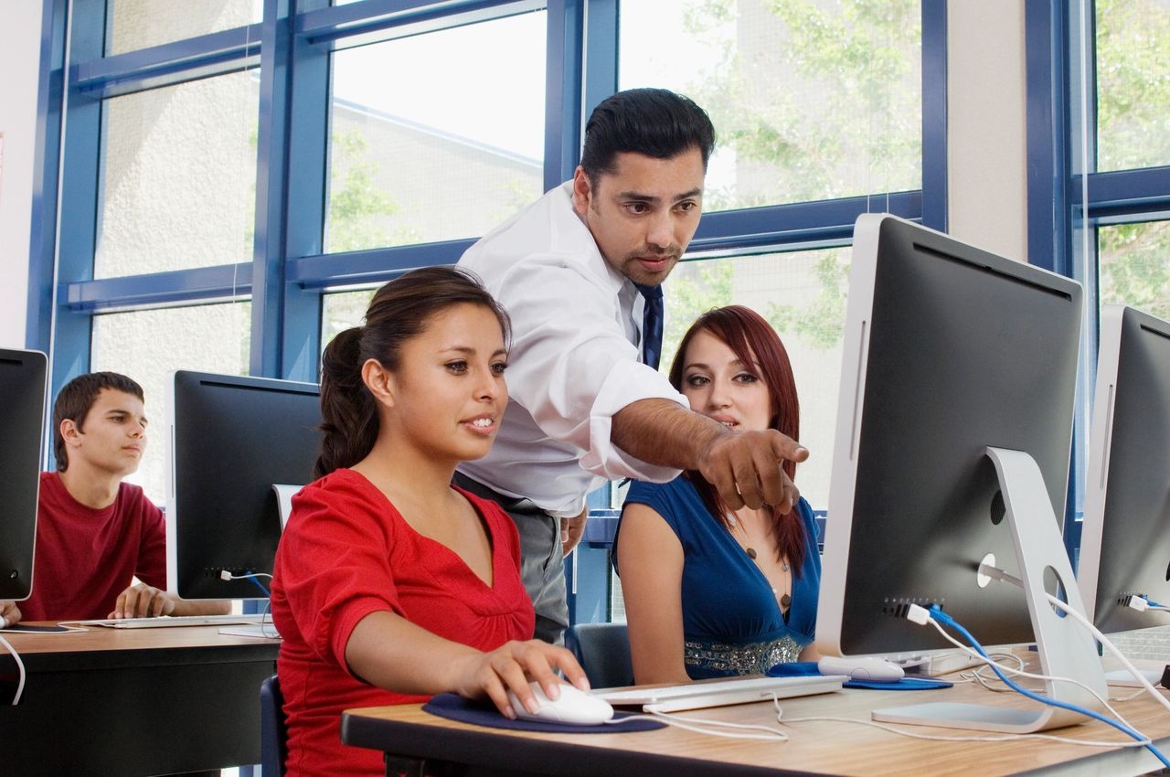 Teacher pointing to the computer screen, showing students data of potential investments.