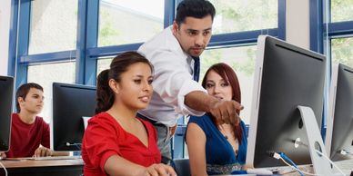 In a classroom, the teacher is pointing to the computer screen of a student