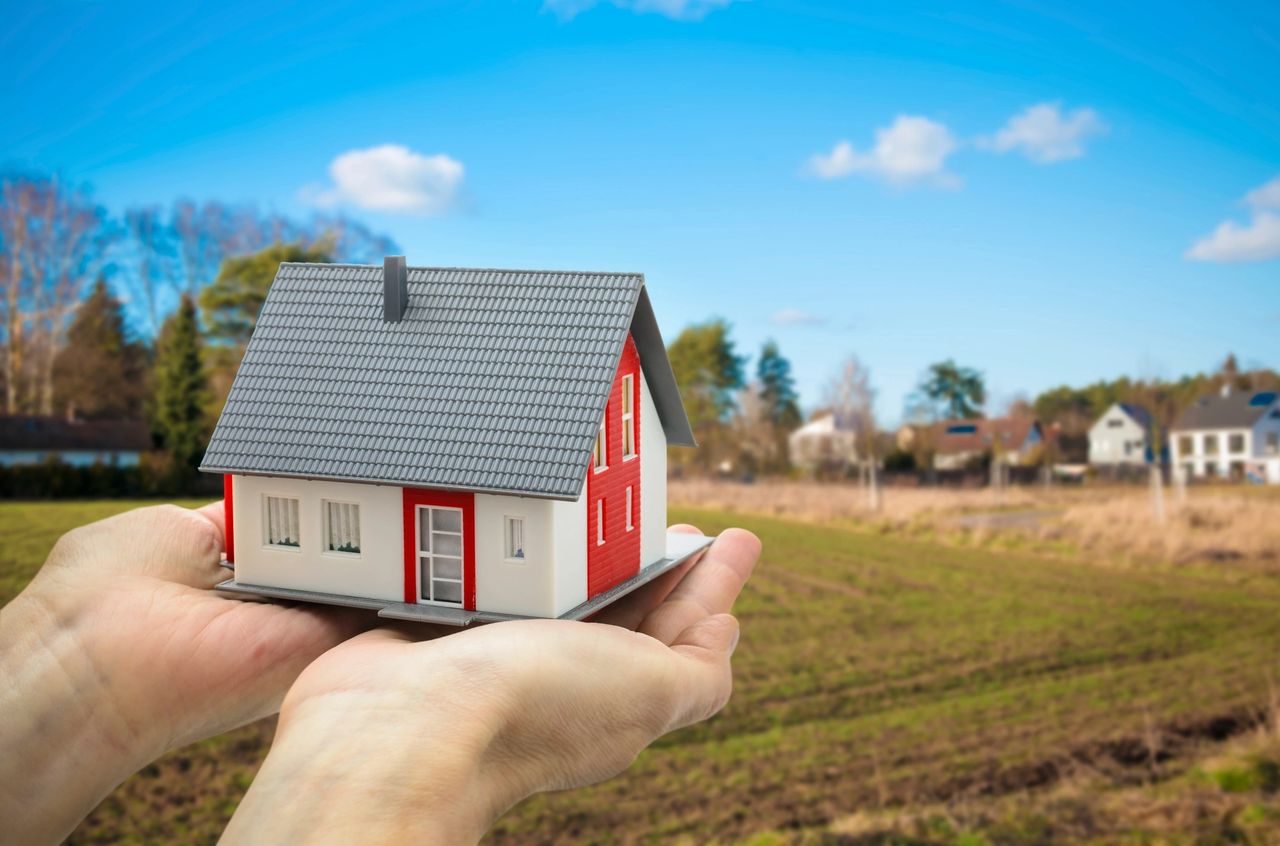 A person holds a home figurine with both hands with a backdrop of an empty lot where the new home will go.